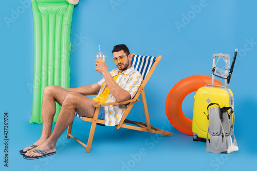 Man in sunglasses holding orange juice on deck chair near inflatable ring and suitcase on blue background © LIGHTFIELD STUDIOS