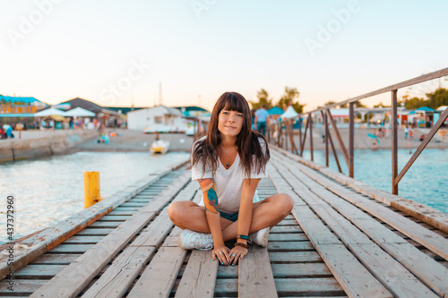 A pretty woman with tattoos sits cross-legged on a wooden pier by the sea. The concept of summer vacation