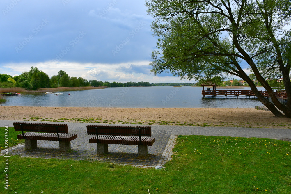 Close up on two wooden benches with concrete bases located next to a sandy beach connected to a vast yet shallow river or lake with some tree visible in the distance together with a wooden marina 