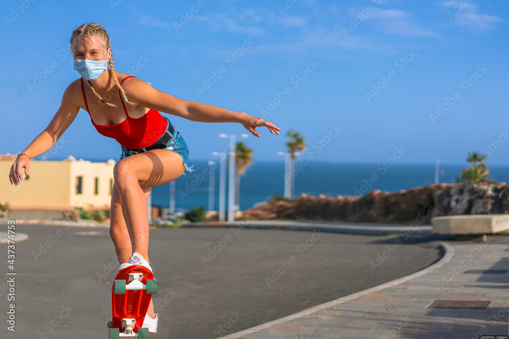 A beautiful teenage girl with blonde hair in a red t shirt and a medical mask skating. Female skater practicing skating on city road. Sport and coronavirus concept.
