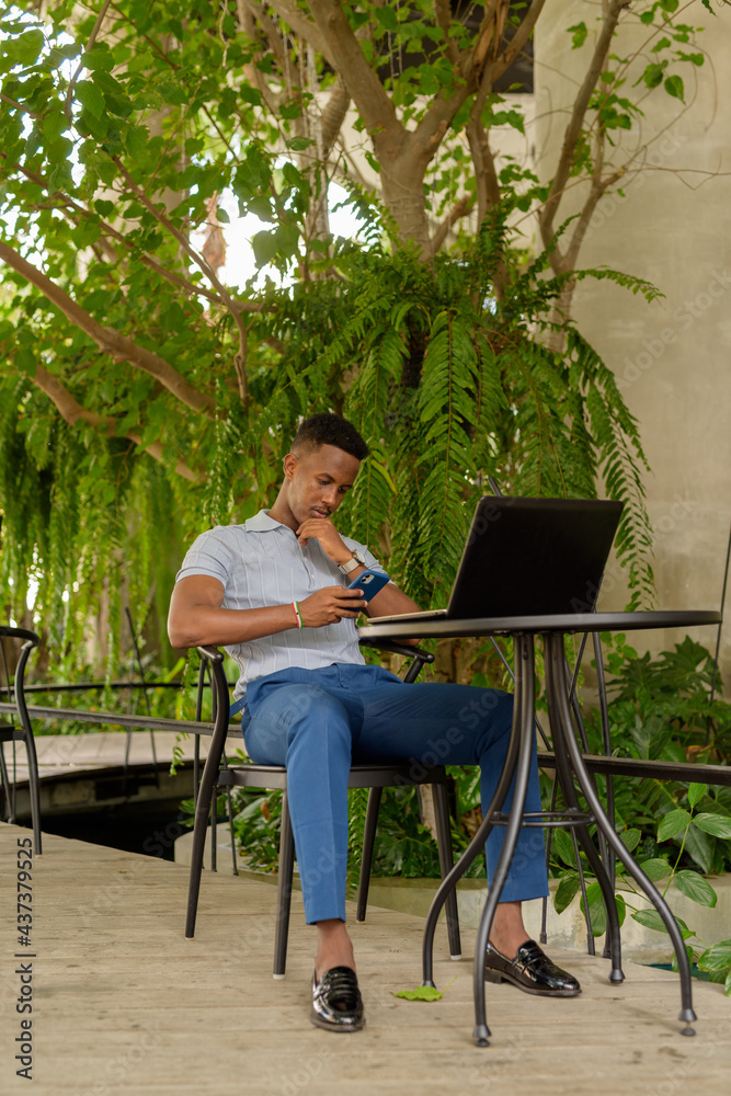 Portrait of young African businessman wearing casual clothes and sitting at coffee shop while using laptop computer and mobile phone while social distancing Portrait of young African businessman wearing casual clothes and sitting at coffee shop while using laptop computer and mobile phone while social distancing