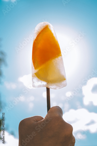 Frozen popsicles on stick in child's hand on background of blue sky. Ice cream with orange and lemon slices, vertical