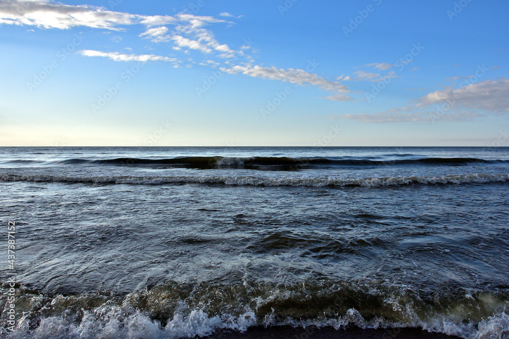 In the seascape, under the blue evening sky with small light clouds, the sea waves roll over the viewer from the front.