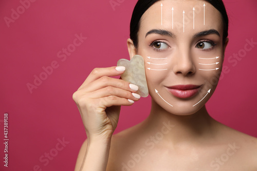 Beautiful young woman doing facial massage with gua sha tool on crimson background, closeup photo