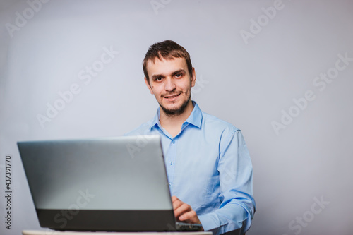 A young handsome man in a blue shirt sits at a computer. Office worker on a white background works for a laptop