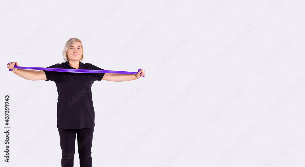 Everyday health for aged people. Sporty senior woman in black sportswear holding rubber resistance band while going to exercise at home. Workout, body goals concept on studio grey background for ad
