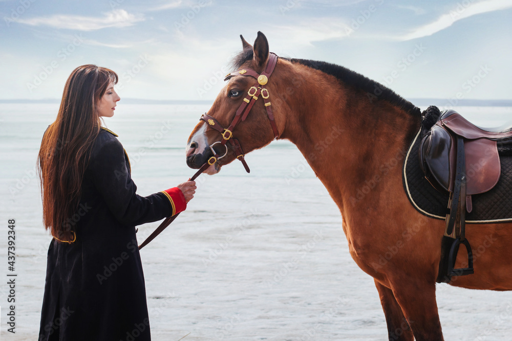 Obraz premium A beautiful woman with long and black hair in a historical hussar costume stands near a river with a horse.
