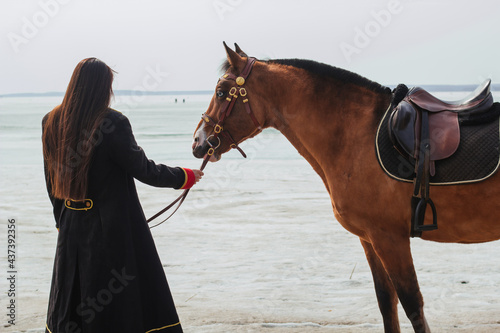 A beautiful woman with long and black hair in a historical hussar costume stands near a river with a horse.