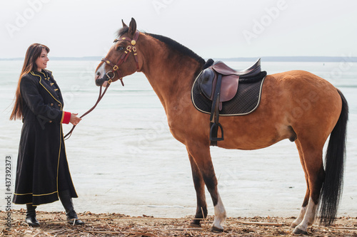A beautiful woman with long and black hair in a historical hussar costume stands near a river with a horse.
