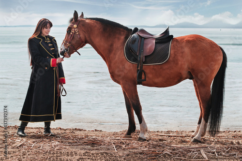 Canvas Print A beautiful woman with long and black hair in a historical hussar costume stands near a river with a horse