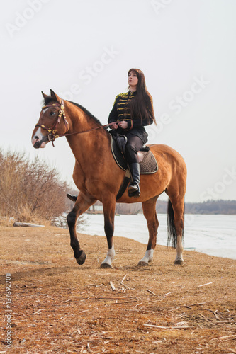 A beautiful woman with long and black hair in a historical hussar costume stands near a river with a horse. © Вероника Преображенс