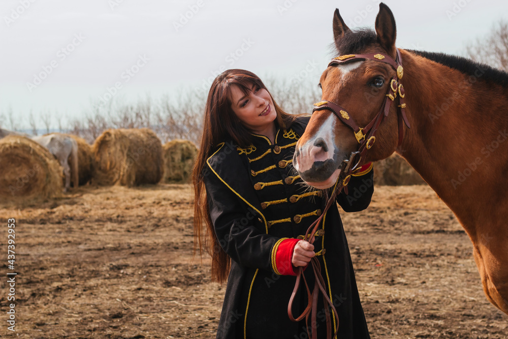 Obraz premium A beautiful woman with long and black hair in a historical hussar costume stands in a field with a horse.