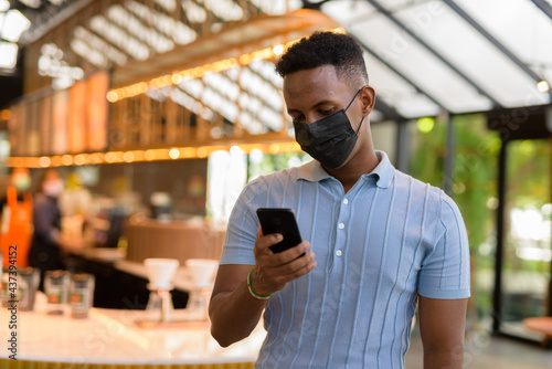 African businessman wearing face mask in coffee shop restaurant while social distancing and using mobile phone