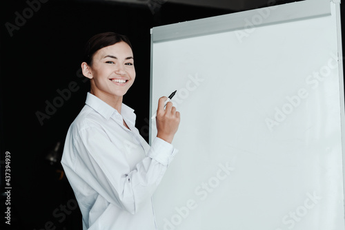 A beautiful girl of Asian appearance, a businesswoman, stands near a white board and holds a felt-tip pen in her hands, smiling