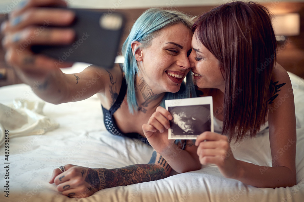 A happy young lesbian couple is lying in the bed full of emotions while taking selfie with ultrasound photo. Home, relationship, love