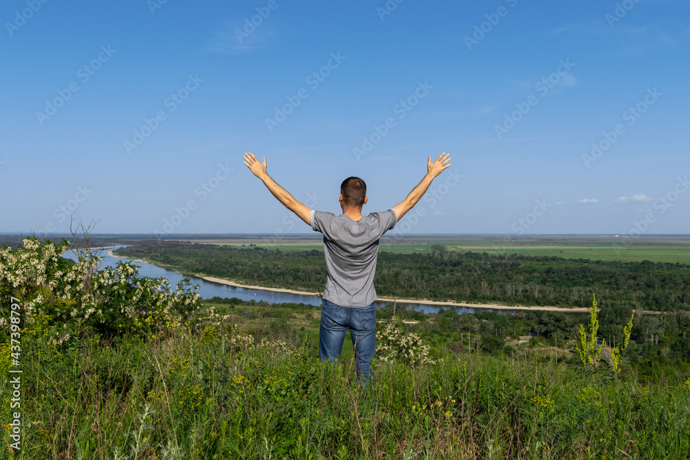 Young man stands on high hill and enjoying natural environment. Happy tourist holds his hands up to the sky and looking at river and green endless fields. Young man stands on high hill and enjoying natural environment. Happy tourist holds his hands up to the sky and looking at river and green endless fields.