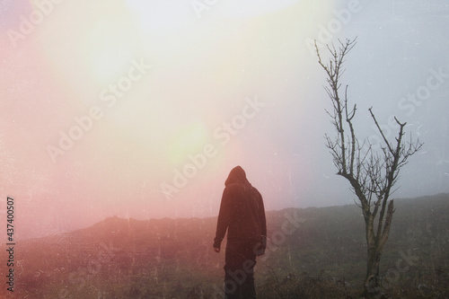 An atmospheric, moody concept. Of a ghostly hooded figure standing by a tree on a foggy day. With a grunge, light leak, retro edit.