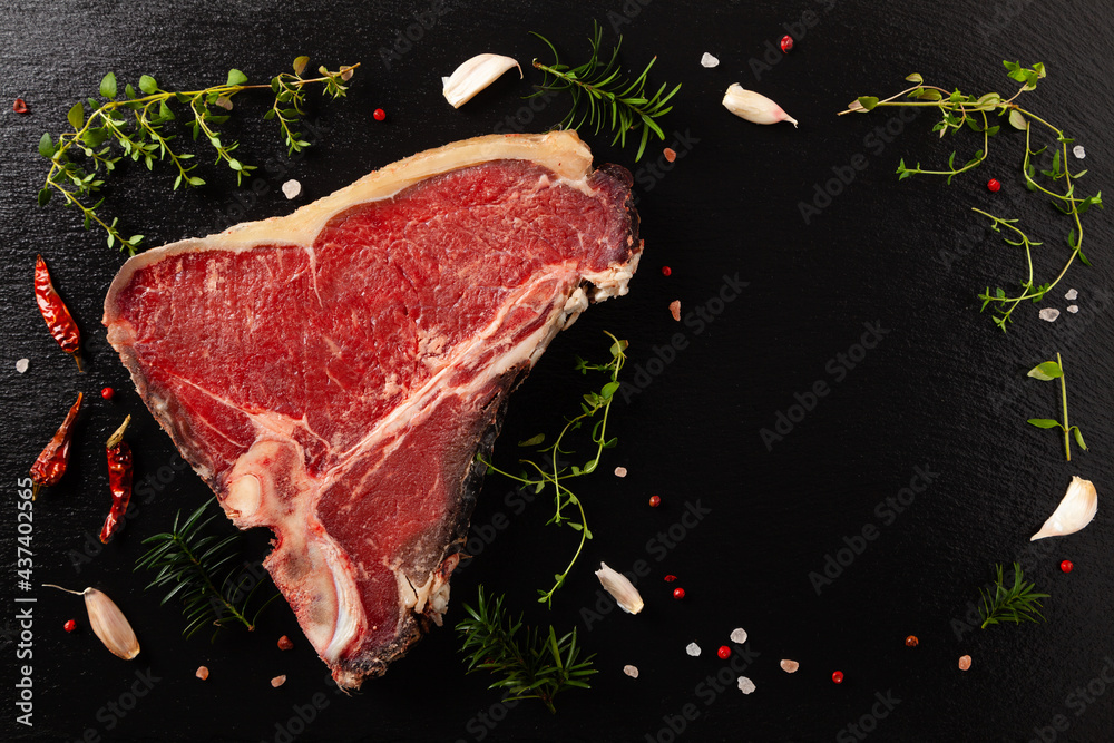 Raw t bone steak, with herbs. Black stone background. View from above.