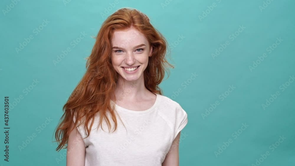 Smiling ginger woman in t-shirt playing with hair and looking at the camera over turquoise background