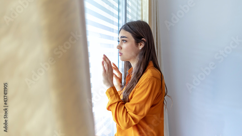 Cropped shot of a beautiful woman looking out the window at home. Sad young woman suffering from agoraphobia looking out of window. Depressed woman standing by window at home. Depressed woman at home. photo