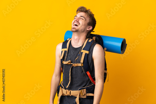 Young mountaineer man with a big backpack isolated on yellow background laughing