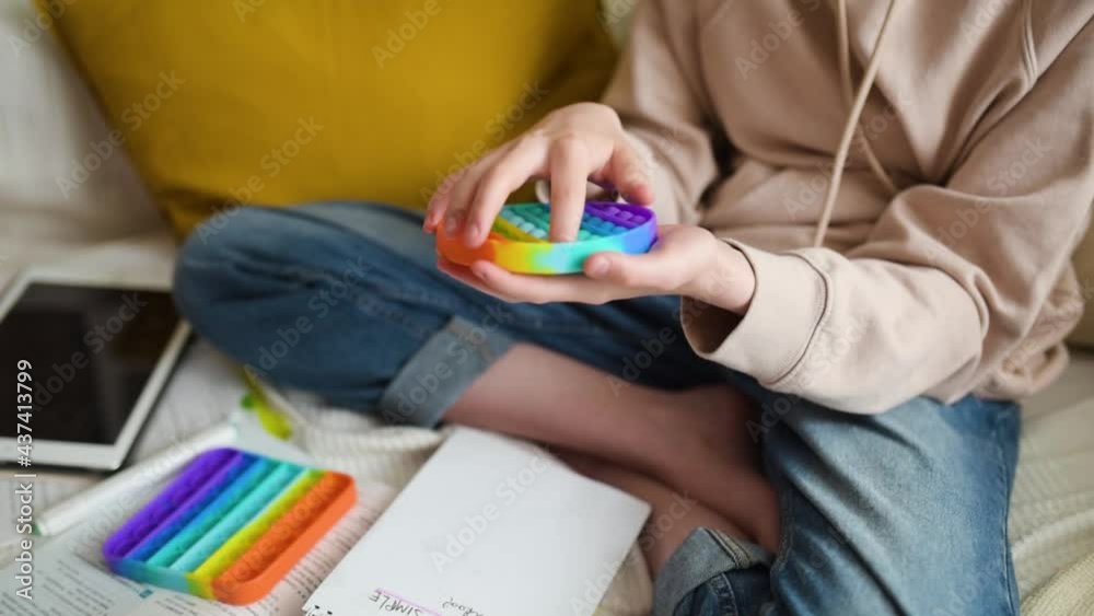Teenage girl playing with rainbow pop-it fidget toy while studying at home. Teen kid with trendy stress and anxiety relief fidgeting game. Popping the dimples of sensory silicone toy.