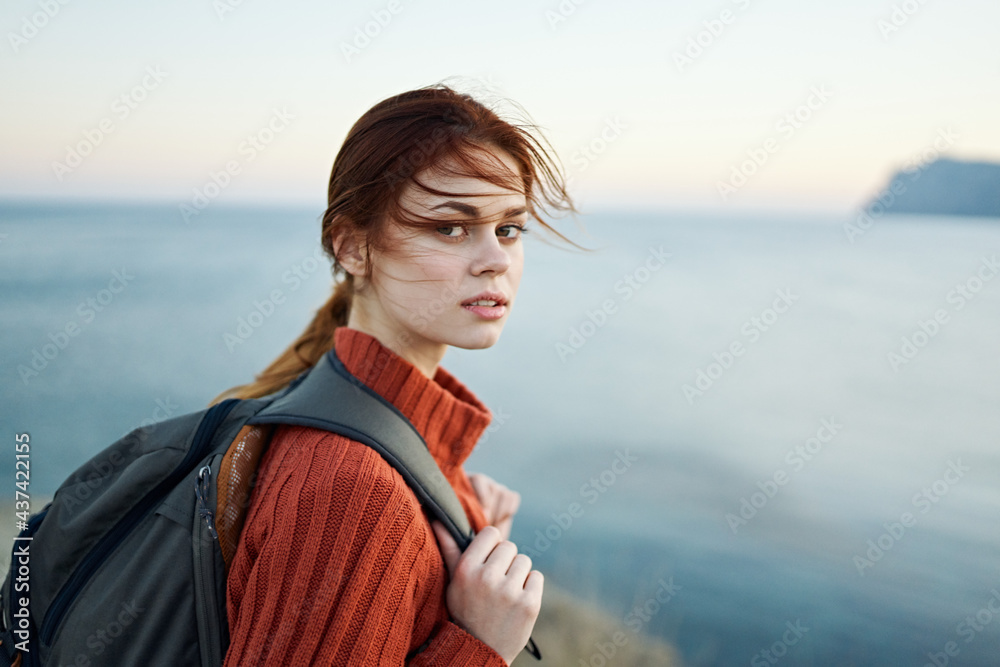 woman tourist in an orange sweater and with a backpack on her back is resting near the sea and mountains in the distance woman tourist in an orange sweater and with a backpack on her back is resting near the sea and mountains in the distance