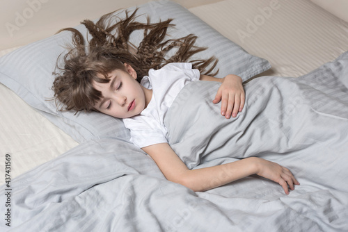 A cute sleeping  little girl with a long wavy hair lies on her back  in a comfort bed on a gray pillow, under a gray blanket on a white sheet. View from above.