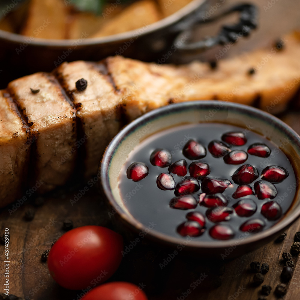 Pomegranate sauce for fried red fish. Sauce bowl with red pomegranate seeds on table with tomatoes and grilled salmon steak on blurred background. Close up shot. Soft focus. Copy space. Pomegranate sauce for fried red fish. Sauce bowl with red pomegranate seeds on table with tomatoes and grilled salmon steak on blurred background. Close up shot. Soft focus. Copy space.