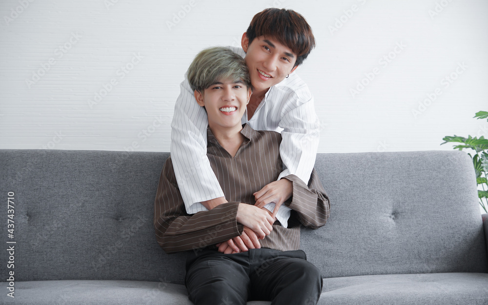 Young attractive LGBT gay couple smiling and hugging from back while they looking at camera and sitting on sofa at home. White background Young attractive LGBT gay couple smiling and hugging from back while they looking at camera and sitting on sofa at home. White background