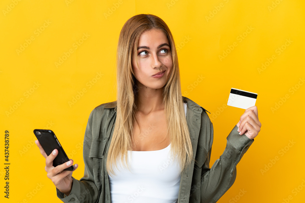 Young blonde woman isolated on yellow background buying with the mobile with a credit card while thinking Young blonde woman isolated on yellow background buying with the mobile with a credit card while thinking