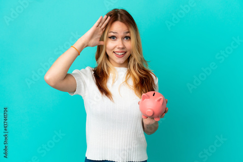 Teenager blonde girl holding a piggybank over isolated blue background with surprise expression