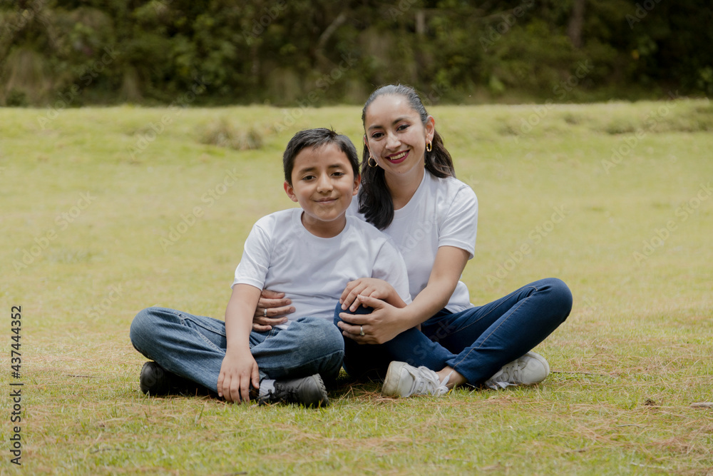 Fototapeta premium Young Hispanic mom with her son sitting in the park-mother hugging her son in the middle of the field outdoors