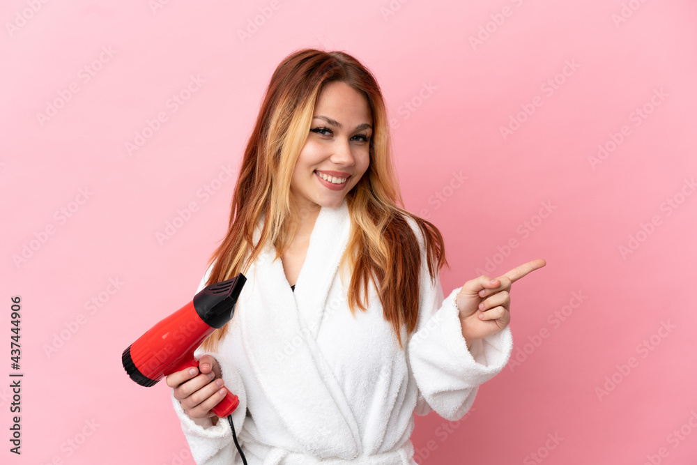 Teenager blonde girl holding a hairdryer over isolated pink background pointing finger to the side Teenager blonde girl holding a hairdryer over isolated pink background pointing finger to the side