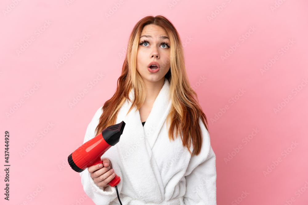 Teenager blonde girl holding a hairdryer over isolated pink background looking up and with surprised expression Teenager blonde girl holding a hairdryer over isolated pink background looking up and with surprised expression