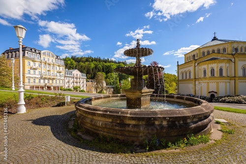 Marianske Lazne, Czech Republic - May 30 2021: View of the baroque water fountain standing on Goethe square. Buildings, church and trees in the background. Sunny day in spa city. Blue sky and clouds. 