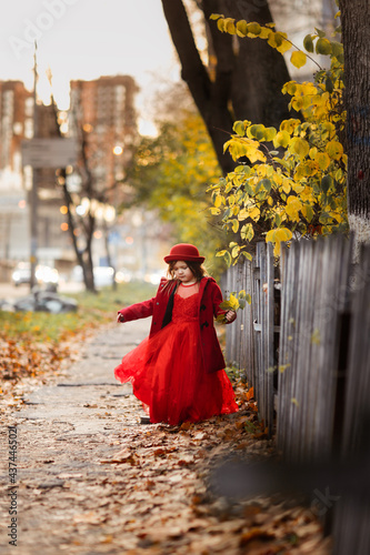 Cute funny caucasian child girl in a red coat walks on the autumn street  stylish child  a girl in a festive dress walking with yellow leaves  autumn mood  a child looks at the leaves of a tree