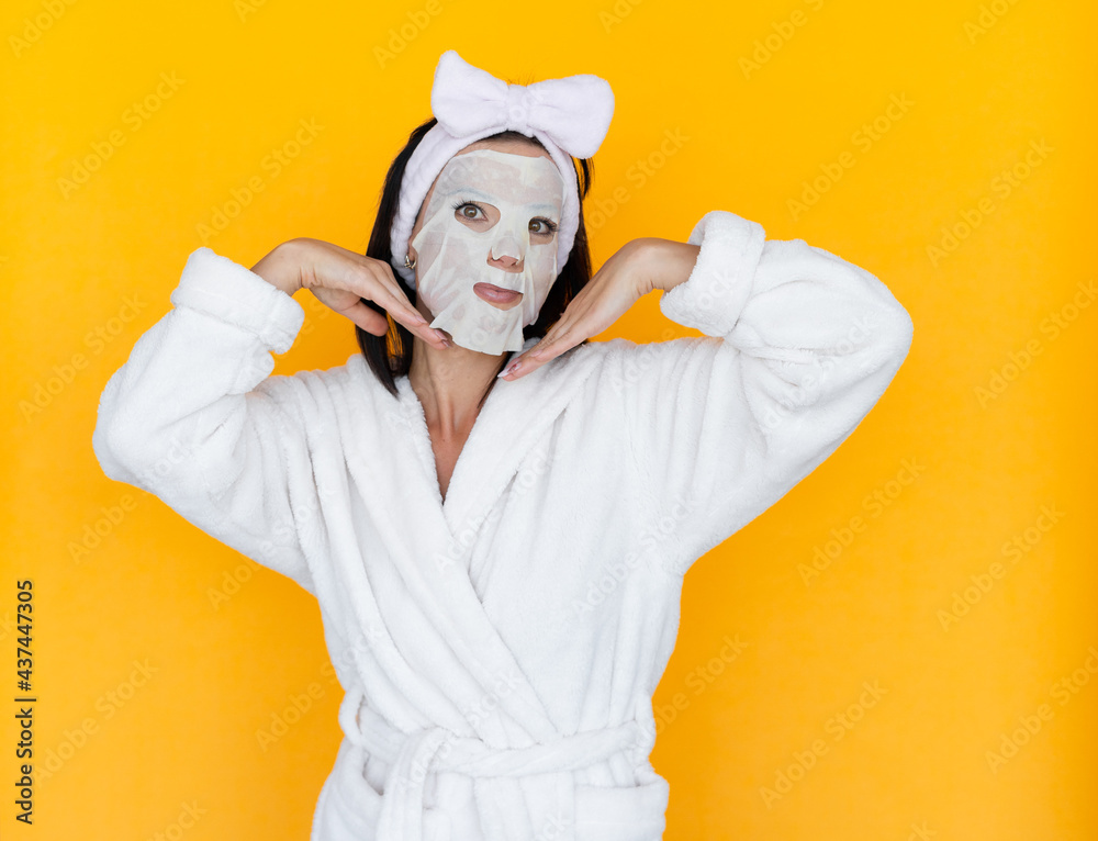 Relax Young woman with tissue facial mask isolated on yellow background, concept for skin care and sunburn protection. Caucasian girl in a white coat standing to the left of the text space.