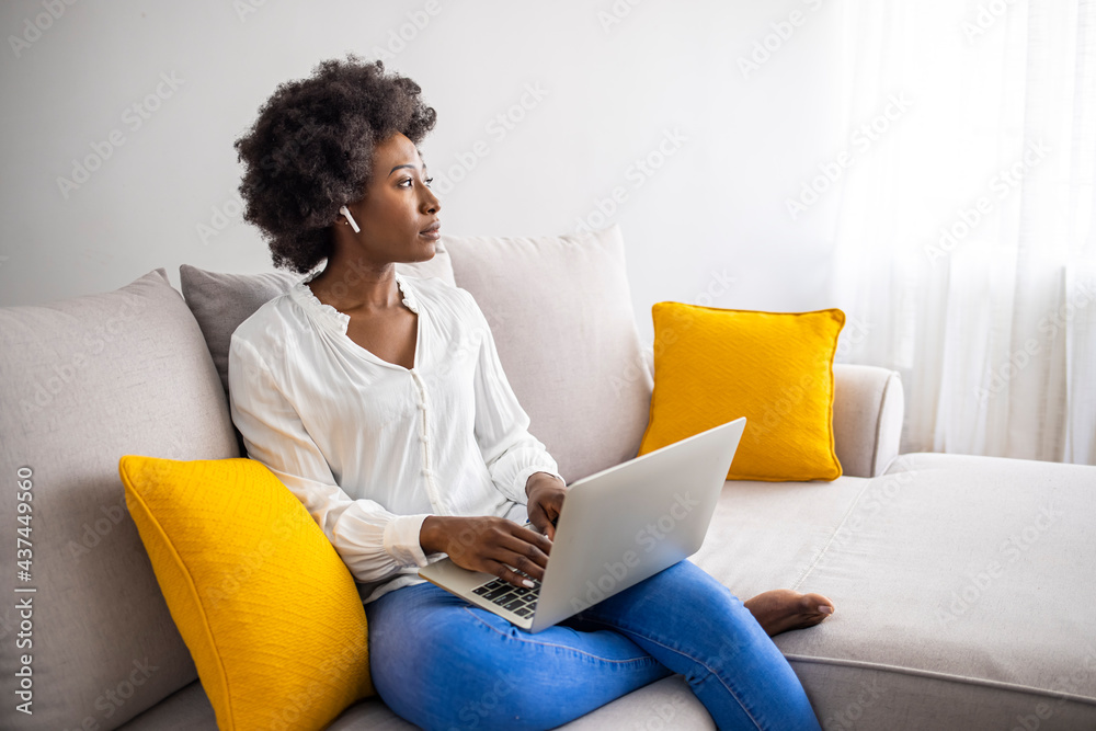 Young woman lying on her living room sofa at home wearing wireless earbuds and working on a laptop. Smiling young woman sitting in her living room at home wearing wireless earbuds and working Young woman lying on her living room sofa at home wearing wireless earbuds and working on a laptop. Smiling young woman sitting in her living room at home wearing wireless earbuds and working