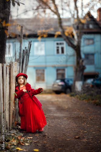 Cute funny Caucasian girl child in a red coat walks on the autumn street near the fence, stylish child, girl in a festive dress for a walk with yellow leaves, autumn mood