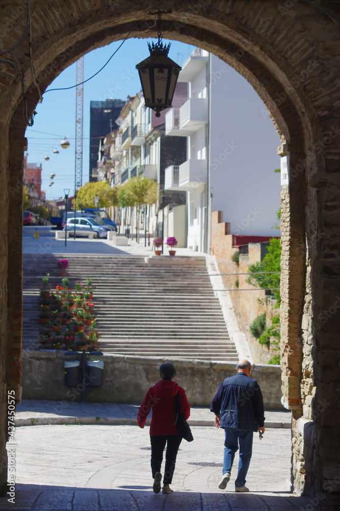 An elderly couple of tourists walk along a stone-paved street passing under an ancient arch where a play of light and shadow emphasizes the scene
