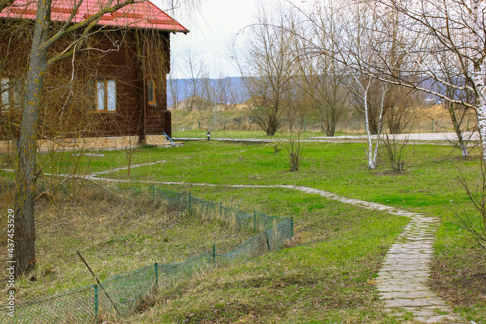 Cobblestone winding road, pathway leading to private home. A wooden modern cottage with red tile roof. Bare trees and green grass on a homestead in the spring, fall season. Private area. Real estate.