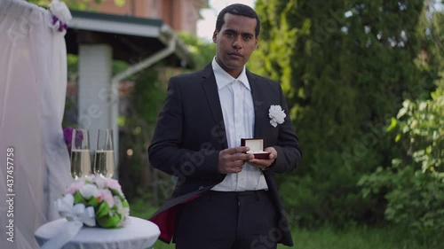 Portrait of African American best man in elegant suit holding ring box looking at camera standing outdoors. Confident handsome young man with jewelry posing at wedding altar in spring garden photo