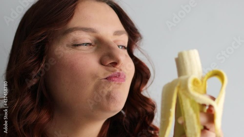 Young woman laughing and eating ripe banana isolated on grey background. Woman biting banana, healthy eating and lifestyle photo