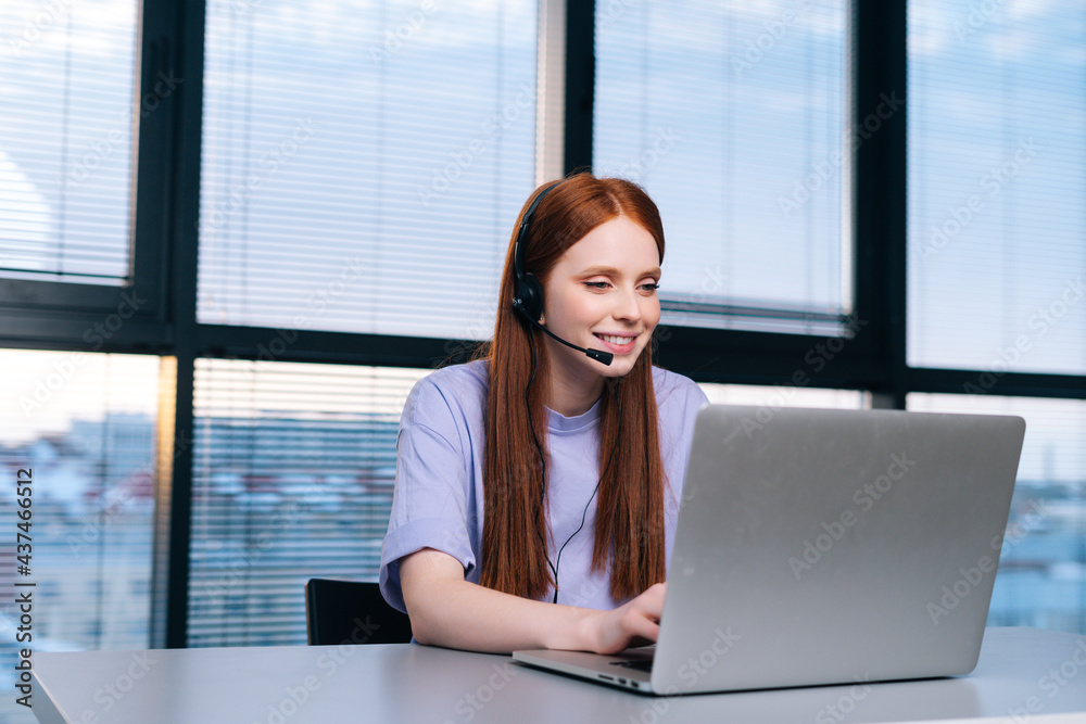Close-up of friendly young woman operator using headset and laptop during customer support at home office. Young female student communicating online by video call on background of window. Close-up of friendly young woman operator using headset and laptop during customer support at home office. Young female student communicating online by video call on background of window.