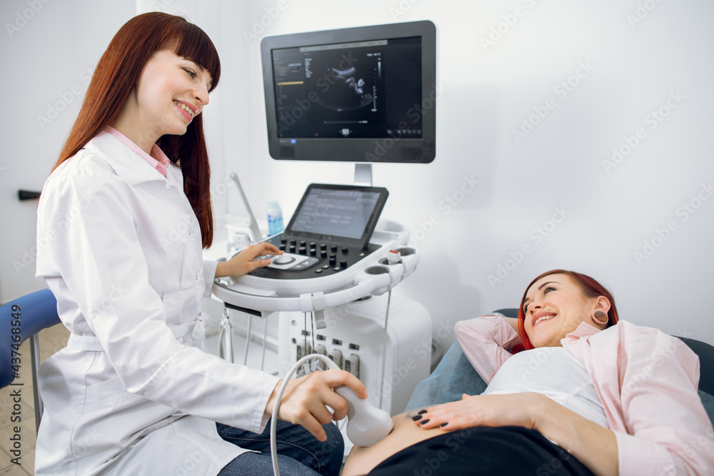 Pretty smiling woman doctor doing an ultrasound diagnostics for her young happy pregnant woman patient. Happy pregnant lady enjoying ultrasound scan of her twins babies Pretty smiling woman doctor doing an ultrasound diagnostics for her young happy pregnant woman patient. Happy pregnant lady enjoying ultrasound scan of her twins babies