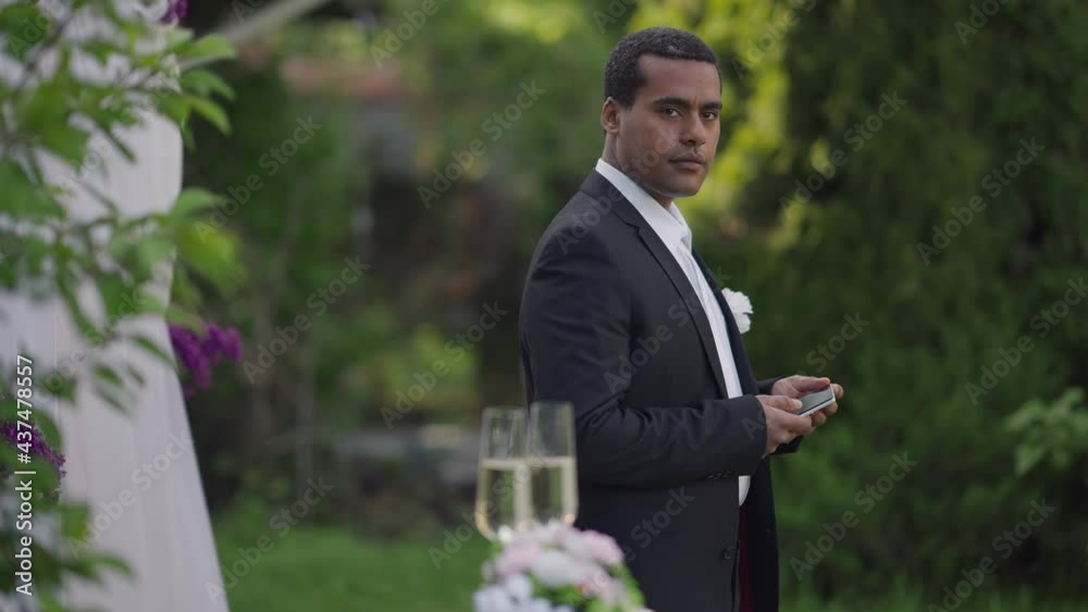 Side view portrait of young groom with smartphone looking around smiling to guests on marriage ceremony. Medium shot of confident elegant man in wedding suit standing at altar outdoors Side view portrait of young groom with smartphone looking around smiling to guests on marriage ceremony. Medium shot of confident elegant man in wedding suit standing at altar outdoors