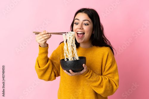 Young caucasian woman isolated on pink background holding a bowl of noodles with chopsticks and eating it photo