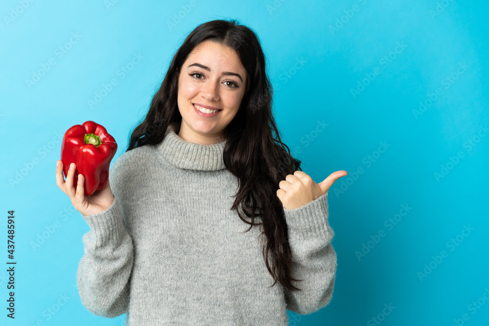 Young caucasian woman holding a pepper isolated on blue background pointing to the side to present a product Young caucasian woman holding a pepper isolated on blue background pointing to the side to present a product