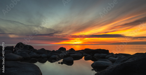 Image of a setting sun with wispy clouds over a calm lake with large dark rocks in the foreground. The clear water reflects the sunset. photo